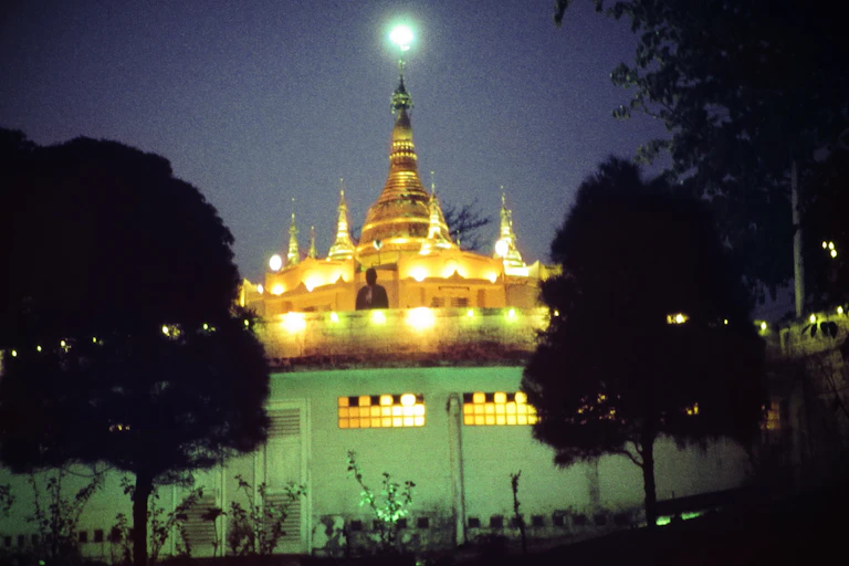 A brightly lit pagoda standing within a green-painted wall, surrounded by trees.