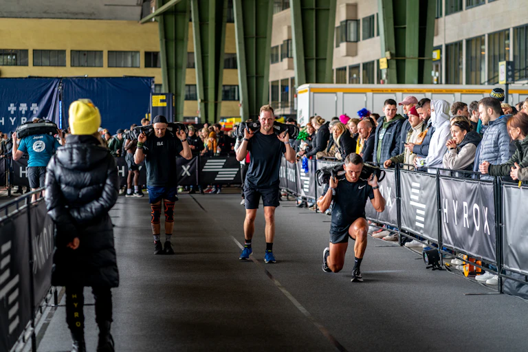 A Hyrox race, depicting a group of men performing lunges with sandbags balanced across their shoulders.