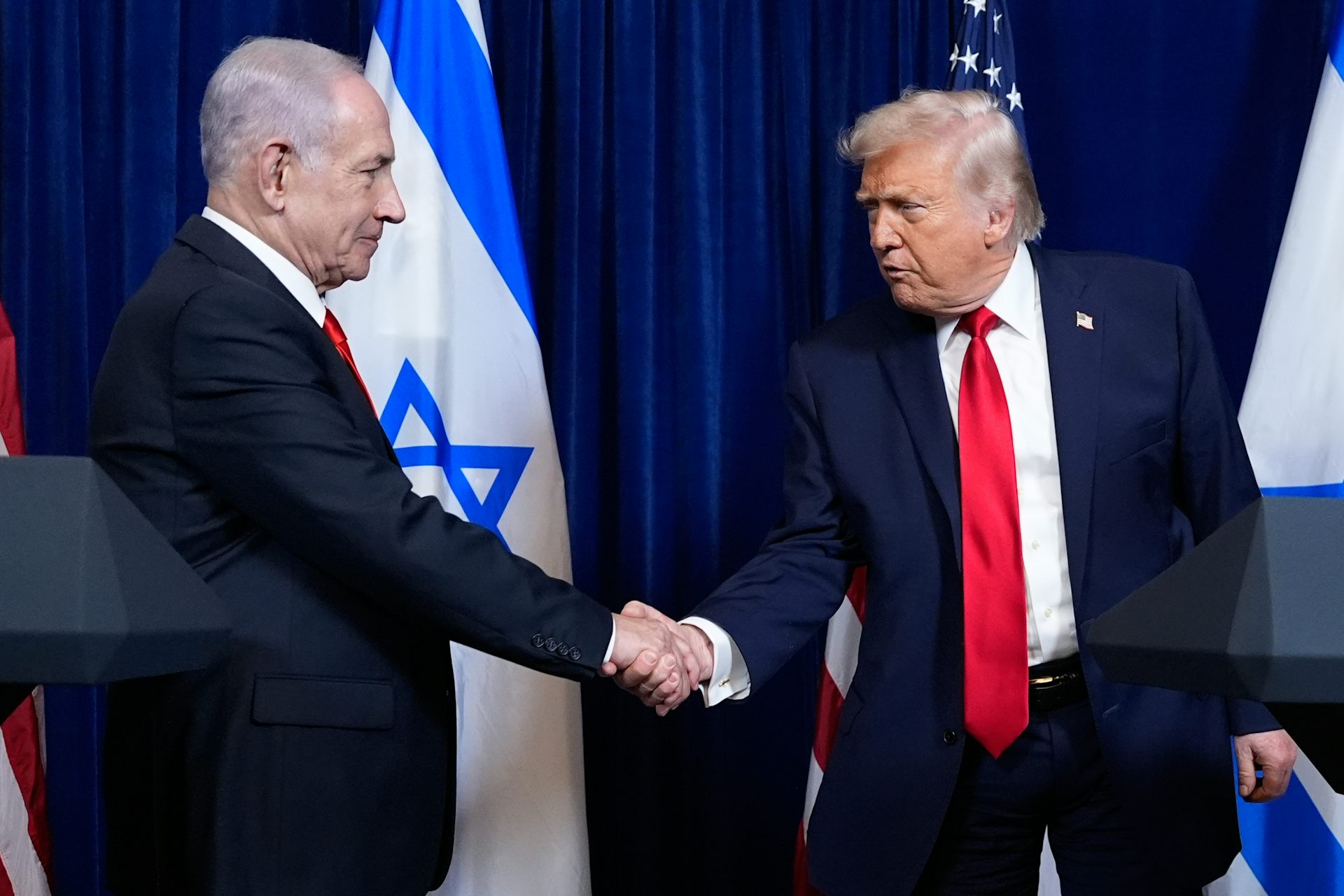Two old white men in suits shake hands in front of the Israeli and American flags