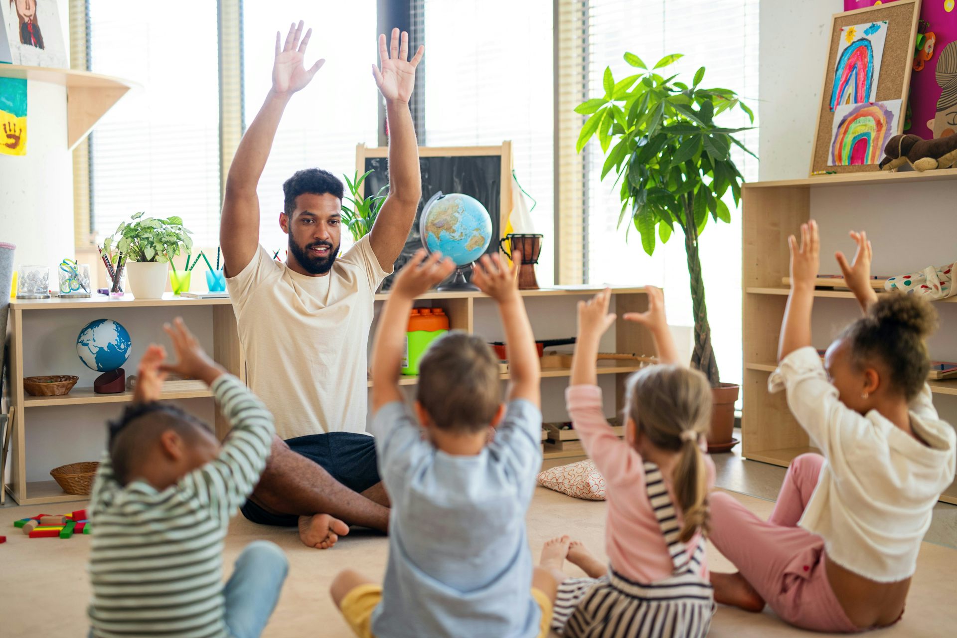 A teacher with his hands up in the air, and children are mirroring him.