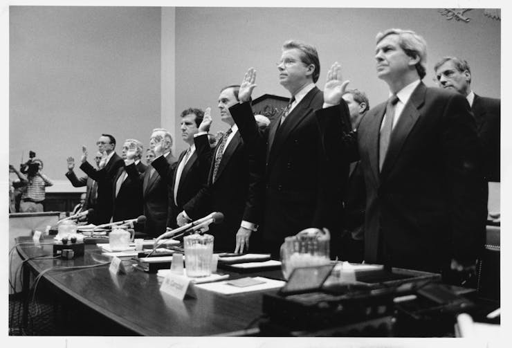 black-and-white photo of eight men in business suits standing behind a table with their right hands raised