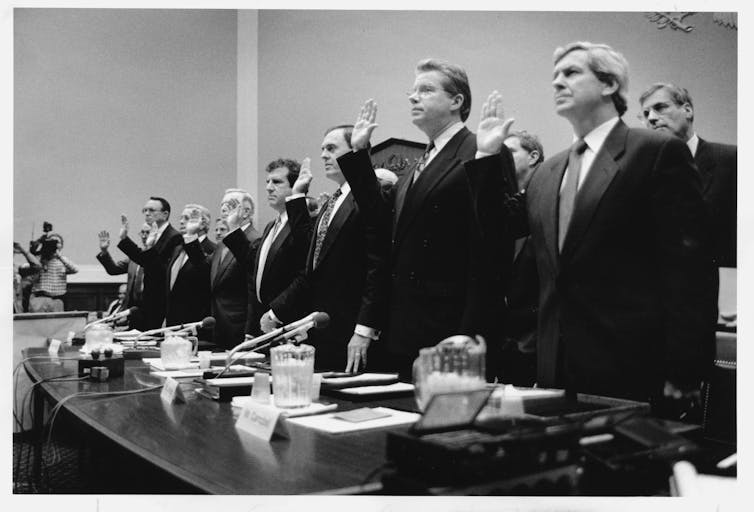 black-and-white photo of eight men in business suits standing behind a table with their right hands raised