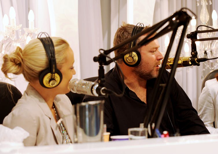 A blonde woman and a man sit at radio microphones wearing over-ear headphones.