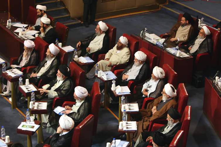 A group of clerics wearing traditional head coverings and long beards sit on seats arranged in concentric rows.