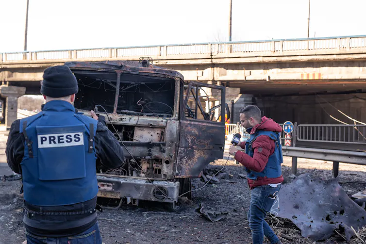 Two male journalists in press flak jackets film around a burned-out truck