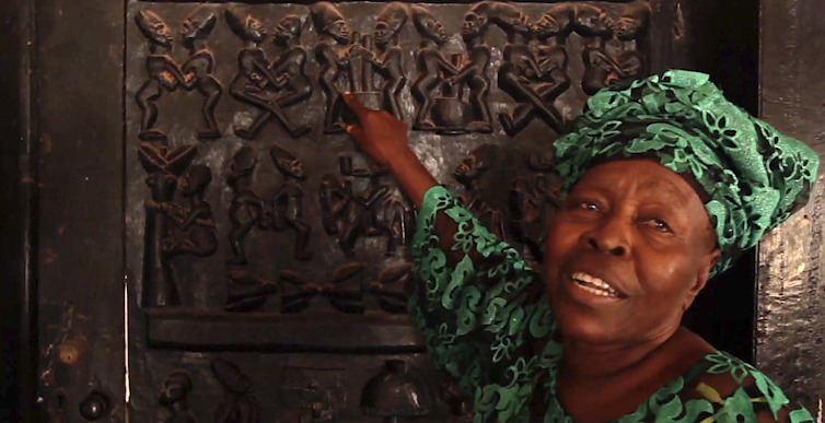 An elder African woman in traditional headgear points to an elaborately carved door.