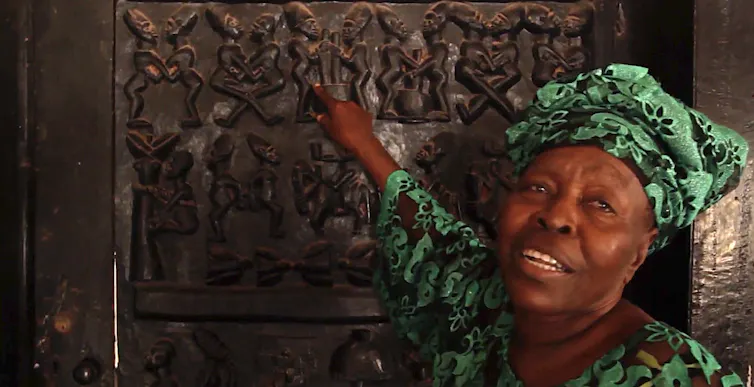 Une femme africaine âgée, coiffée d'un couvre-chef traditionnel, montre du doigt une porte finement sculptée.