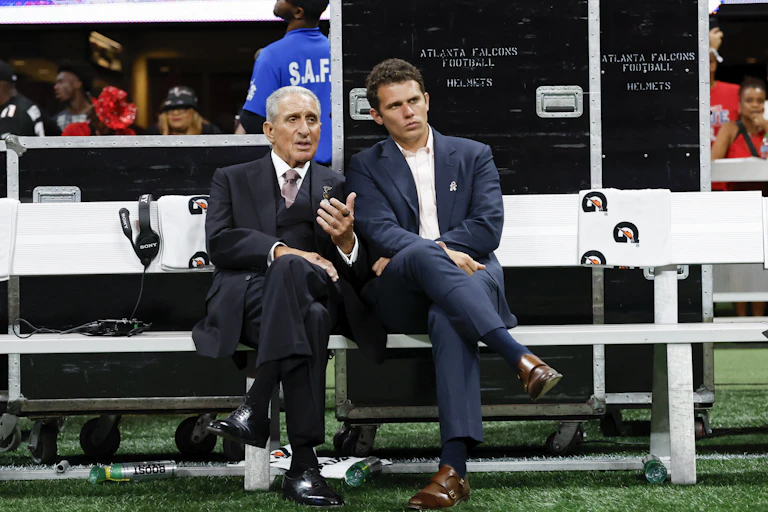 Two men in suits talk while sitting on a bench with athletes milling around in the background.
