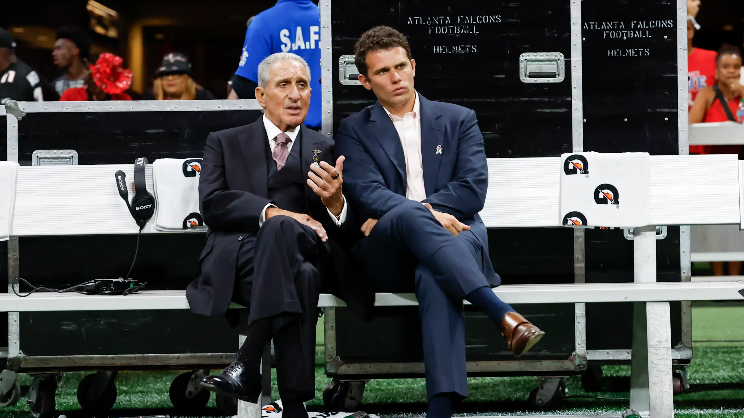 Two men in suits talk while sitting on a bench with athletes milling around in the background.