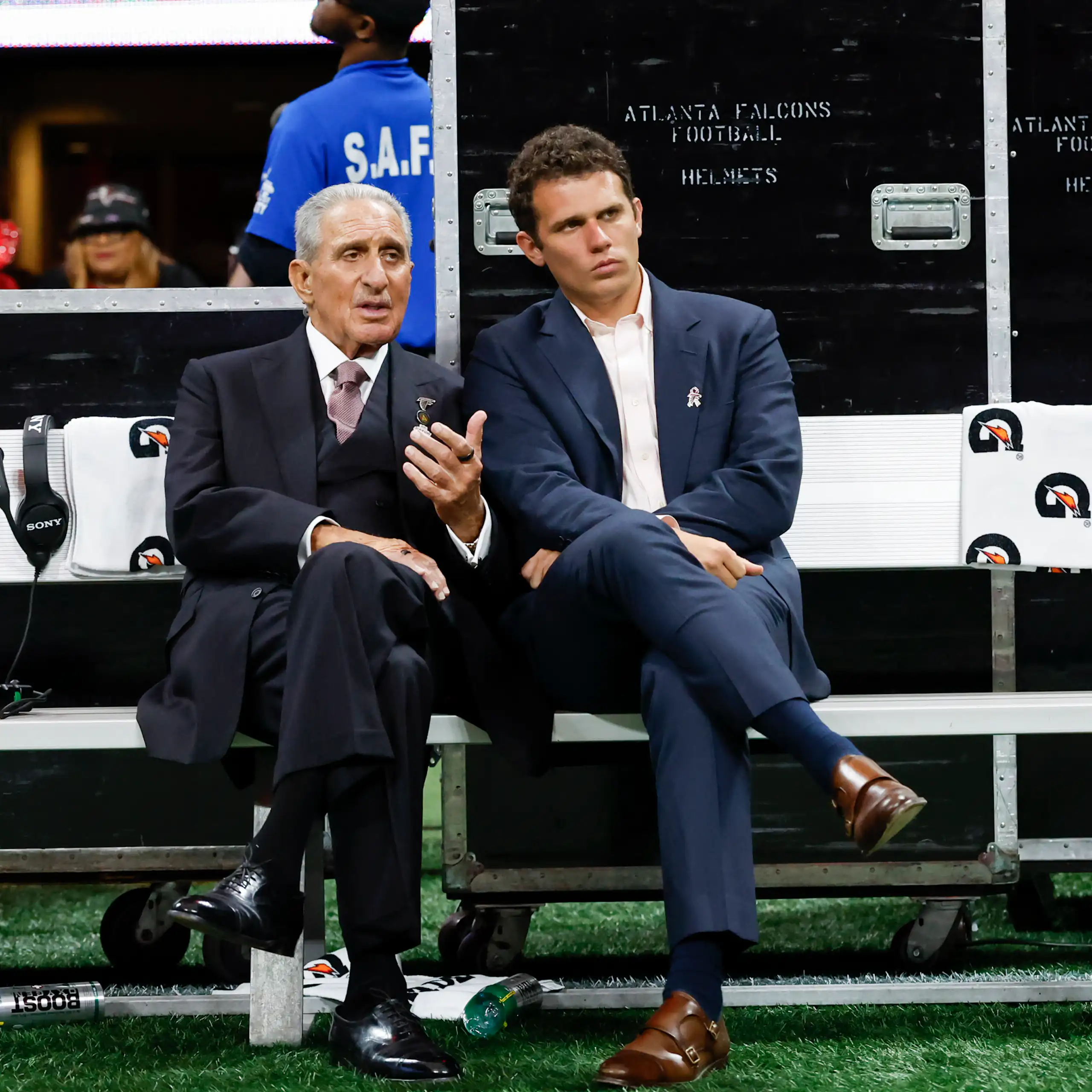 Two men in suits talk while sitting on a bench with athletes milling around in the background.
