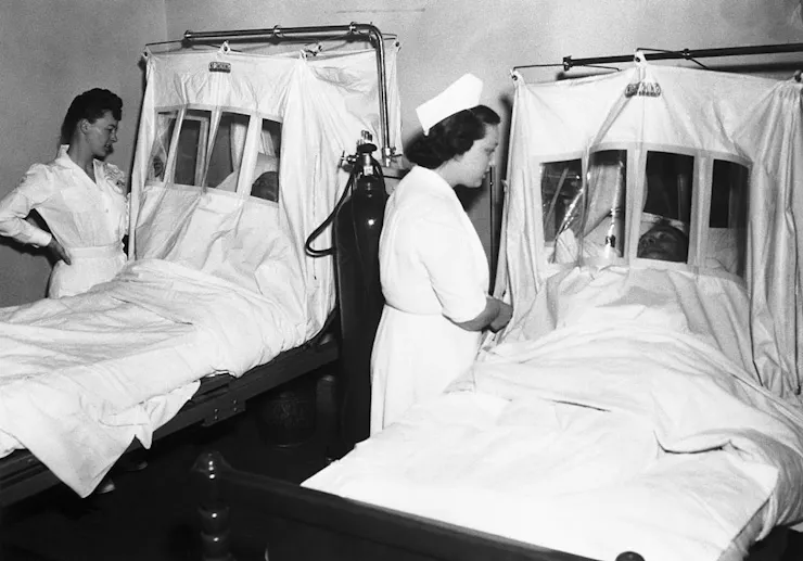 In a old black and white photo, two nurses administer oxygen to patients in tented hospital beds.
