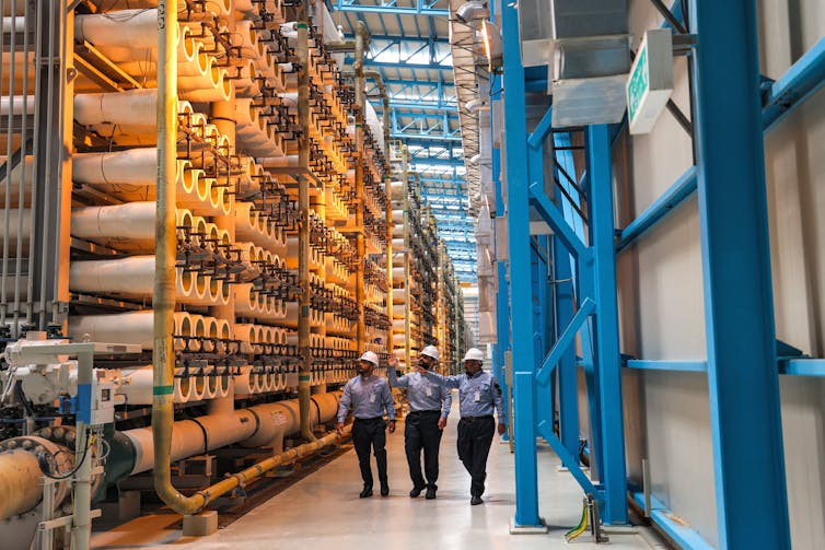 Three people walk through a massive space with many large pipes and valves.