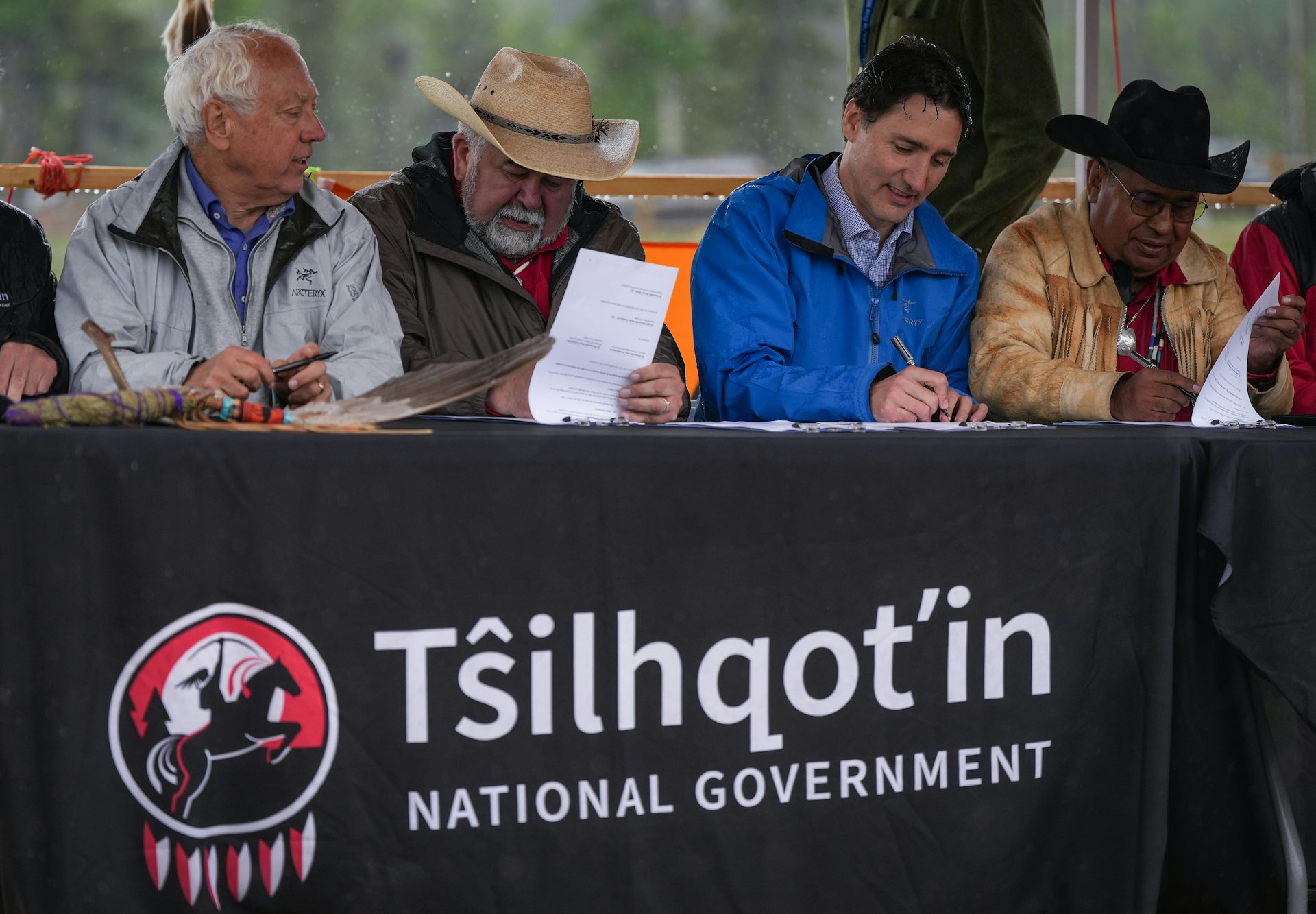 Un hombre de pelo oscuro con una cazadora azul firma un documento en una mesa con otros tres hombres, dos de los cuales llevan sombreros de vaquero.