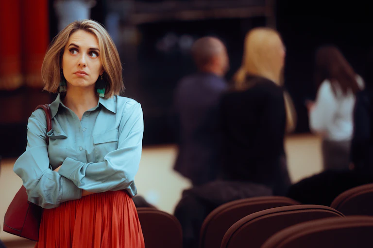 introverted young woman standing alone at a social event with other people in the background.