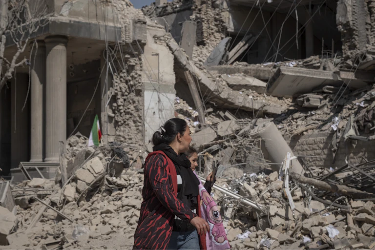 A woman wearing a red jumper and a young girl carrying a pink blanket walk past a pile of grey rubble.