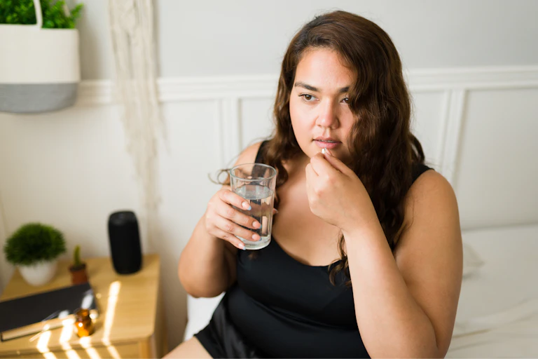 A woman holds a glass of water in one hand and a pill in the other had, which she goes to place in her mouth.