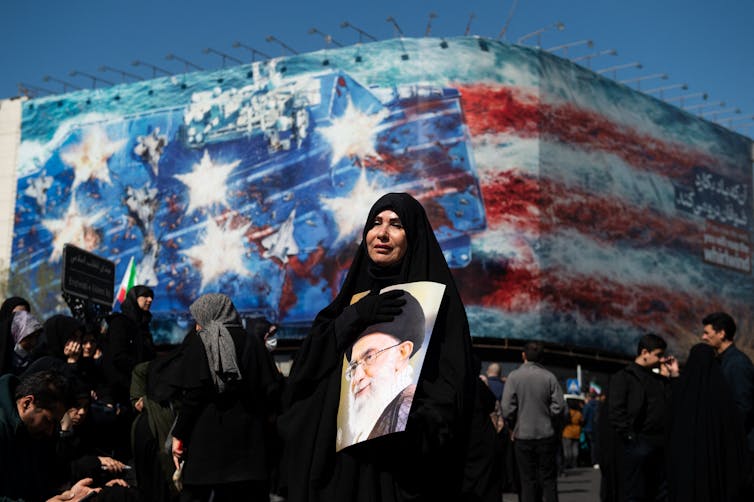 A woman in a black headcovering holding a large photo of a man with a white beard, glasses and wearing a black turban.