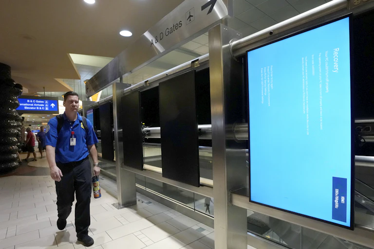 a man wearing an ID lanyared and carrying a water bottle walks past blank display screensin an airport terminal