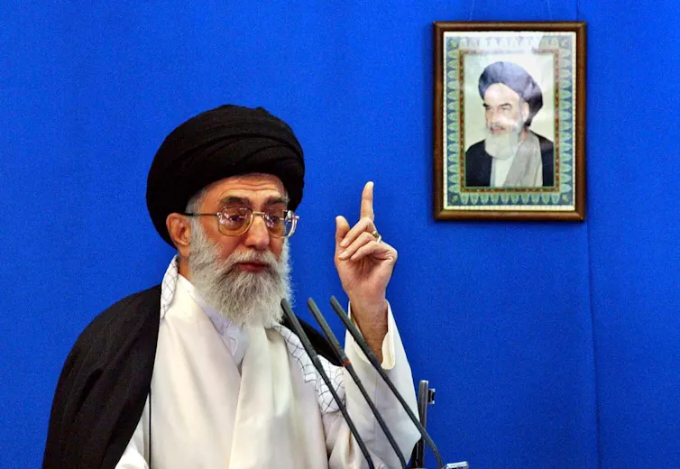 A bearded cleric in a black turban, talking in front of a framed photo of a different cleric.