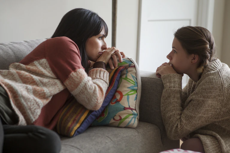 Woman lying on couch and adolescent girl leaning on it look at each other