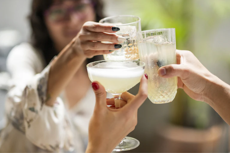 A young woman in the background holds up a glass to two others' drinks in a 'cheers.'