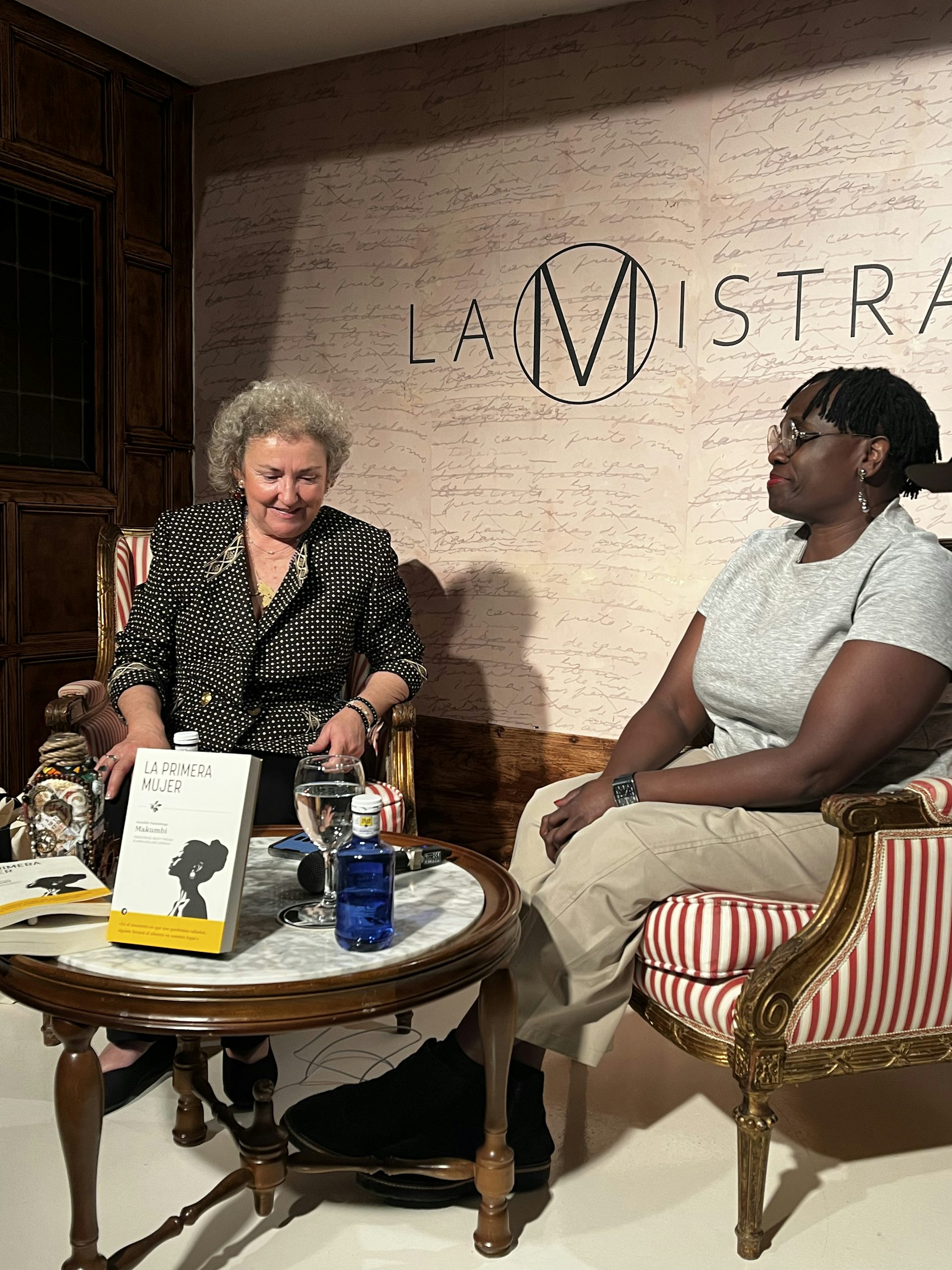 Dos mujeres, una blanca y otra negra, sentadas en una mesa baja mientras conversan en la presentación de un libro.