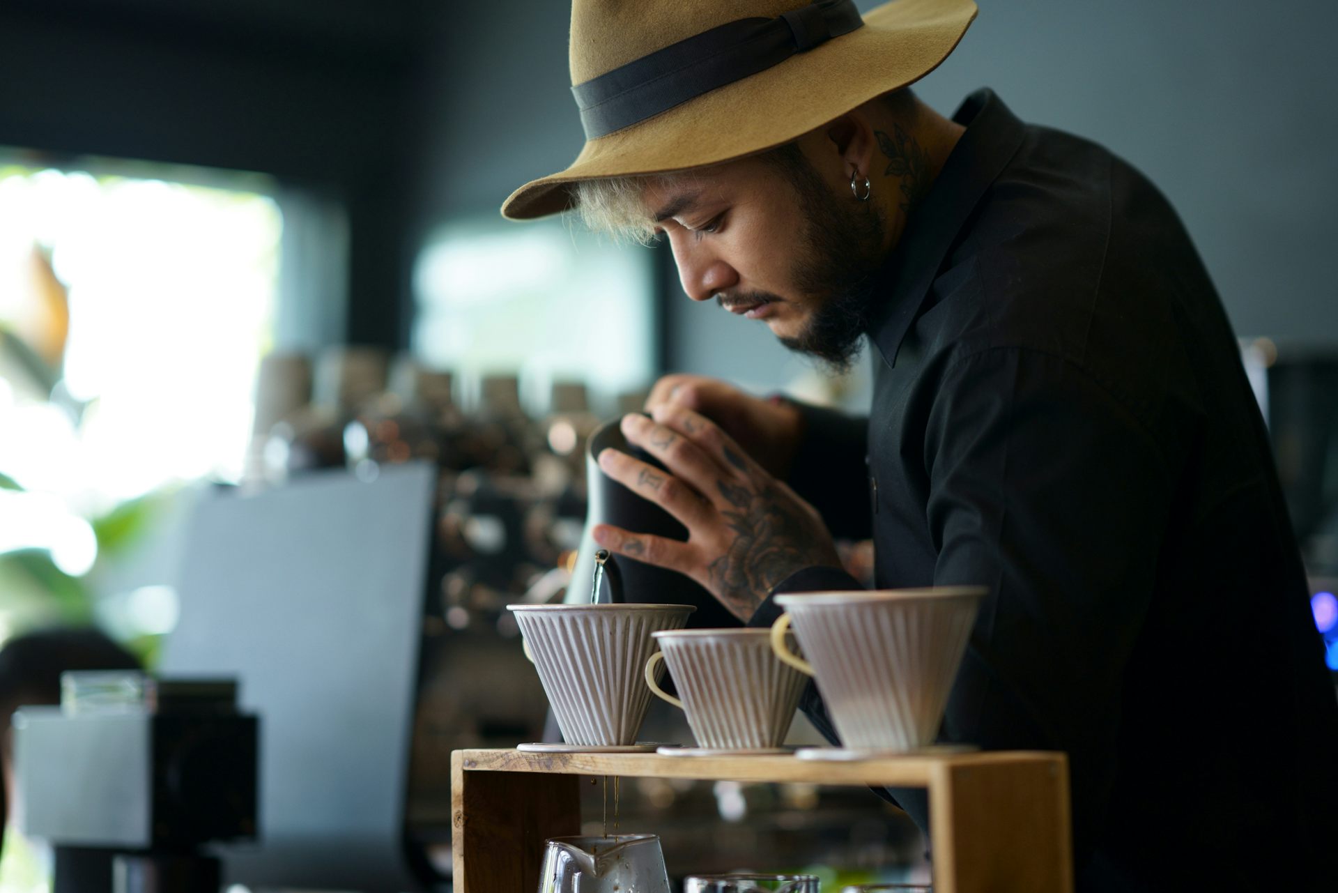 Barista with a beard and tattooed hands pours boiling water over coffee grounds.