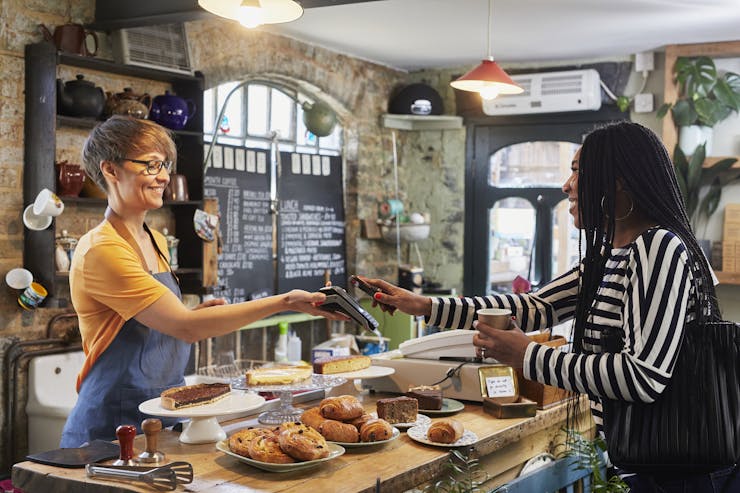 A young woman with dreadlocks pays for her coffee as a smiling young female barista with short hair holds out a card reader.