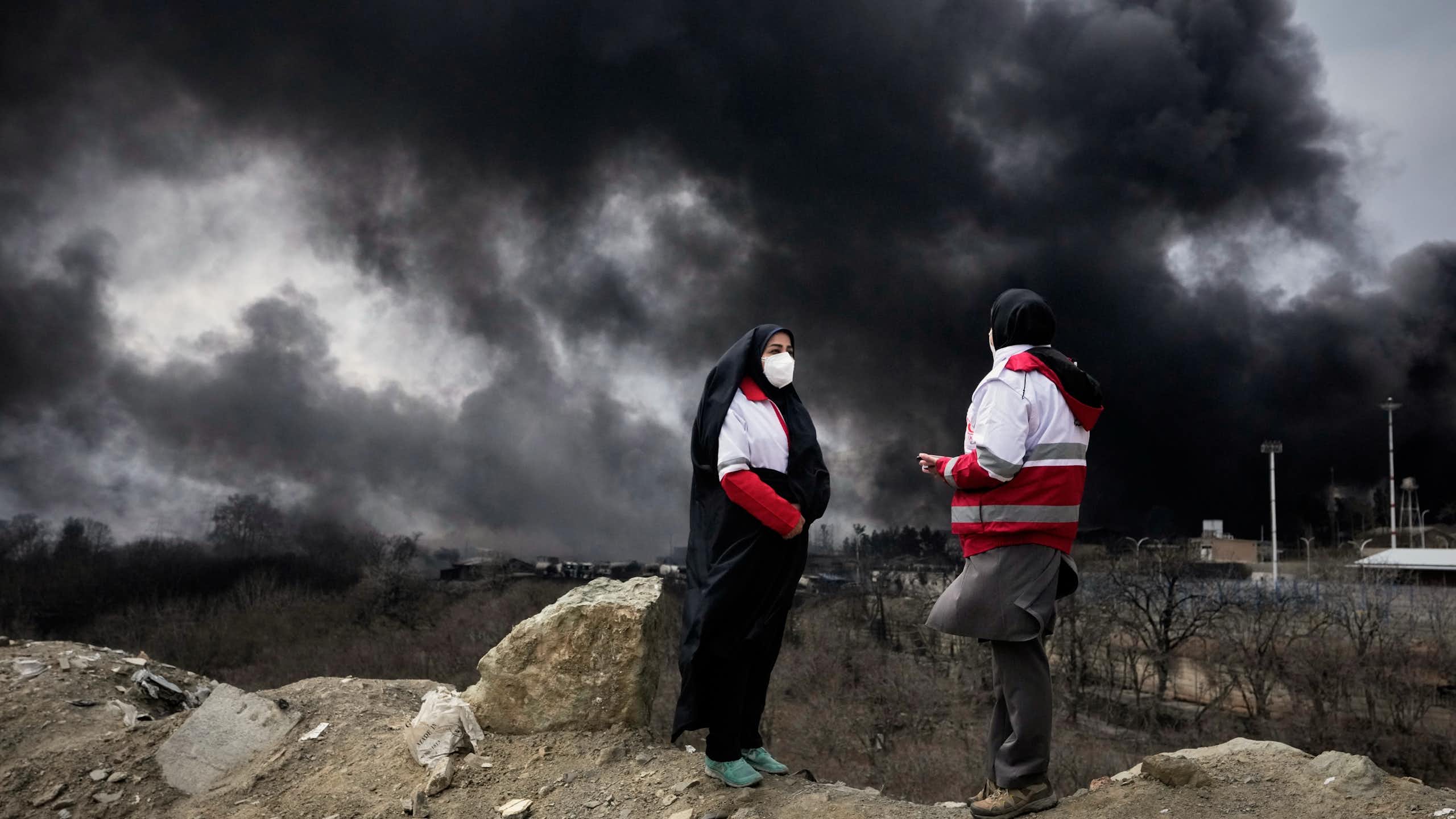 Two women standing outdoors with thick black plumes of smoke in the sky