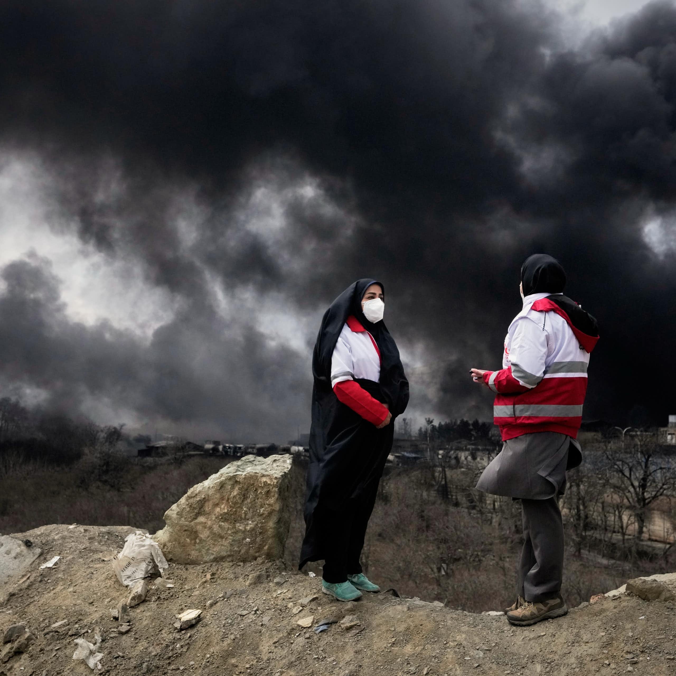 Two women standing outdoors with thick black plumes of smoke in the sky