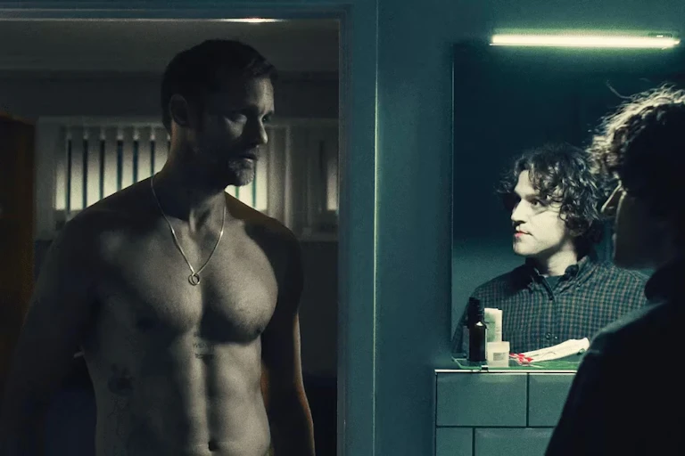 A shirtless man gazes at a man wearing a shirt and the dressed man is also seen in the reflection of a mirror.