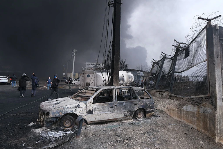 A bombed out white car in front of a fenced area, with a black plume of smoke rising in the background and a handful of people wearing face masks walking in the area.
