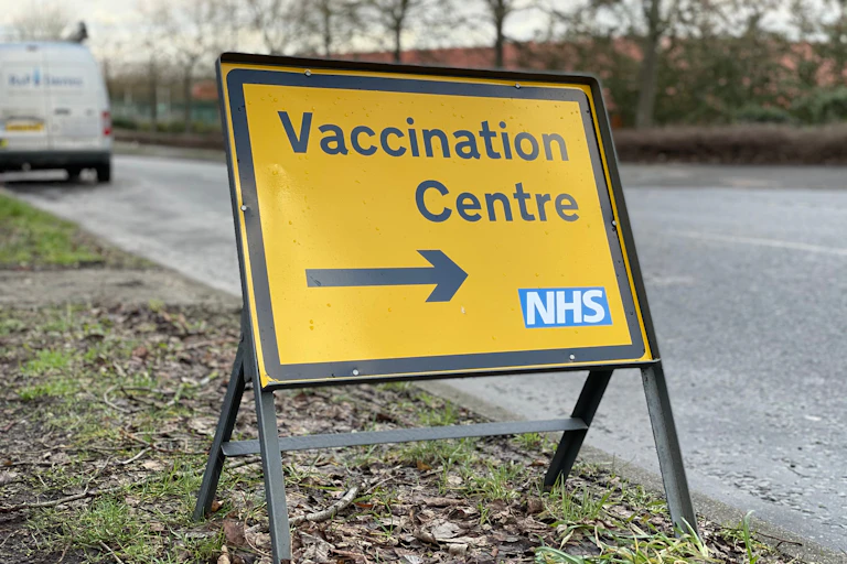 A yellow sign at the side of the road pointing to an NHS vaccination centre.