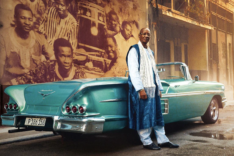 An African man leans against a classic turquoise convertible in a street with a mural of musicians behind him.