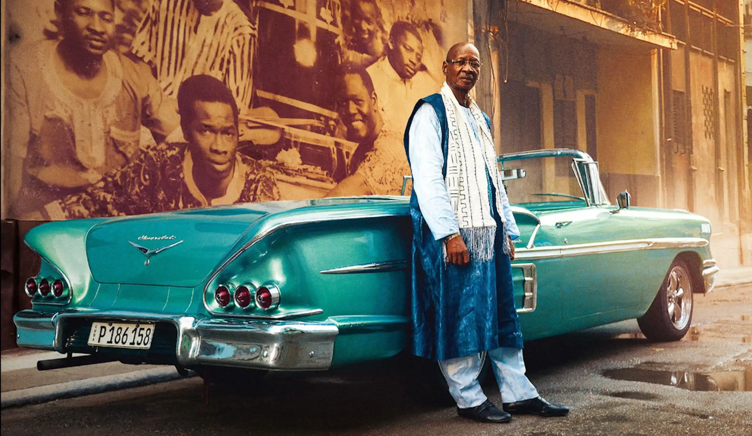 An African man leans against a classic turquoise convertible in a street with a mural of musicians behind him.