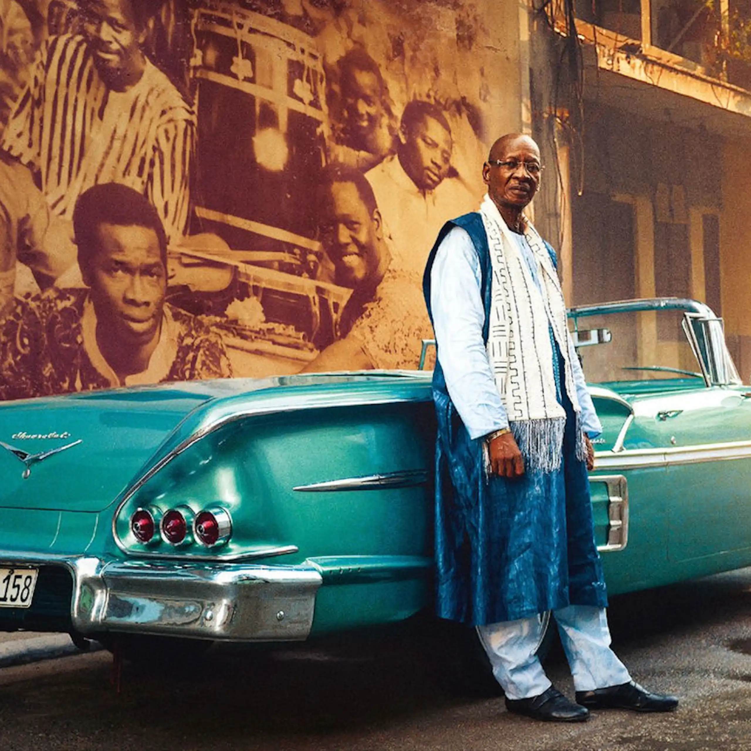 An African man leans against a classic turquoise convertible in a street with a mural of musicians behind him.