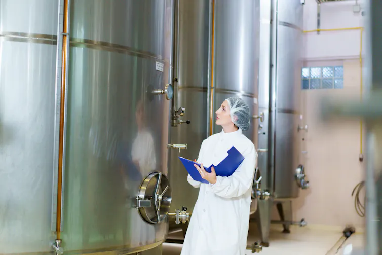 woman in white lab coat and holding clipboard looks at big metal vat inside factory