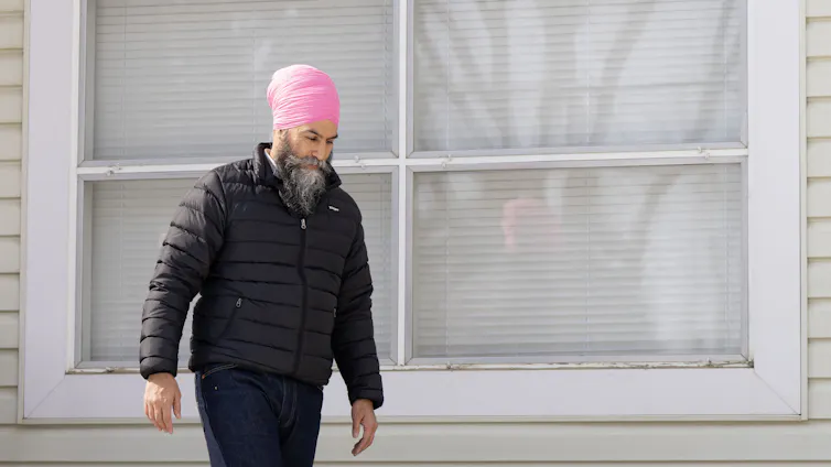 A bearded man in a pink turban walks past a house.
