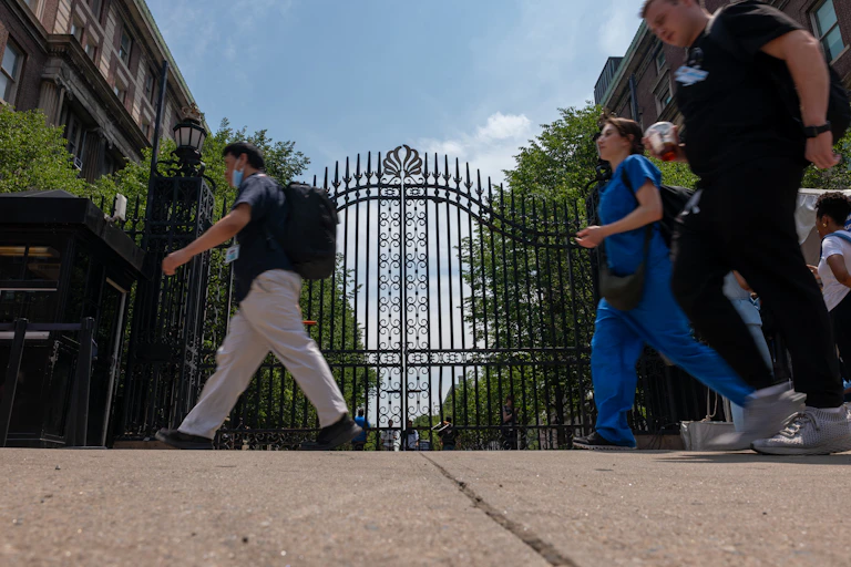Several people walk past large gates on a sidewalk.
