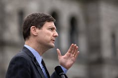 Un hombre con traje oscuro habla al aire libre con la mano levantada, visto de perfil.