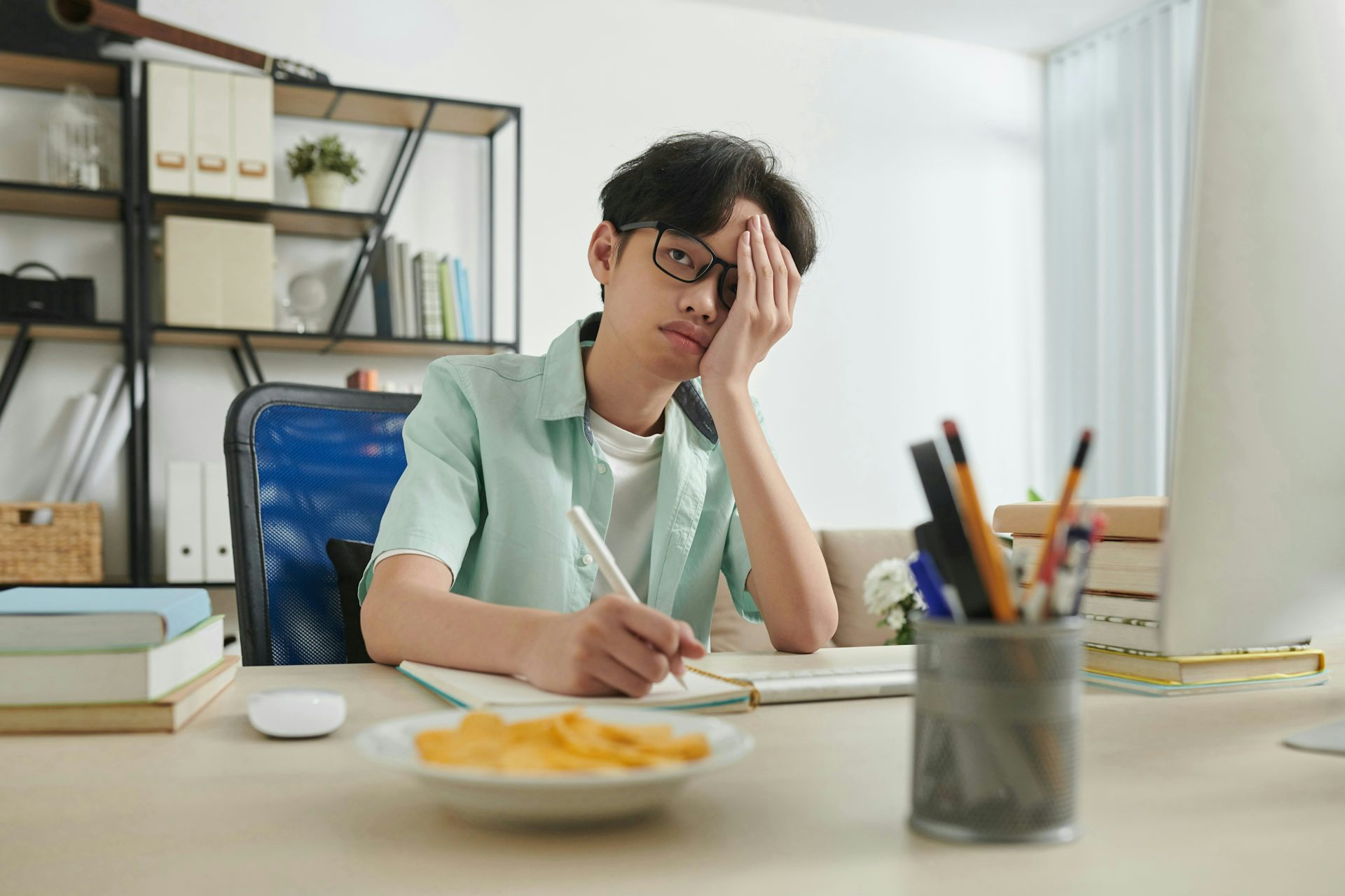 Un adolescente con gafas se sienta en un escritorio con un cuaderno y un bolígrafo, apoyando la cara en la mano