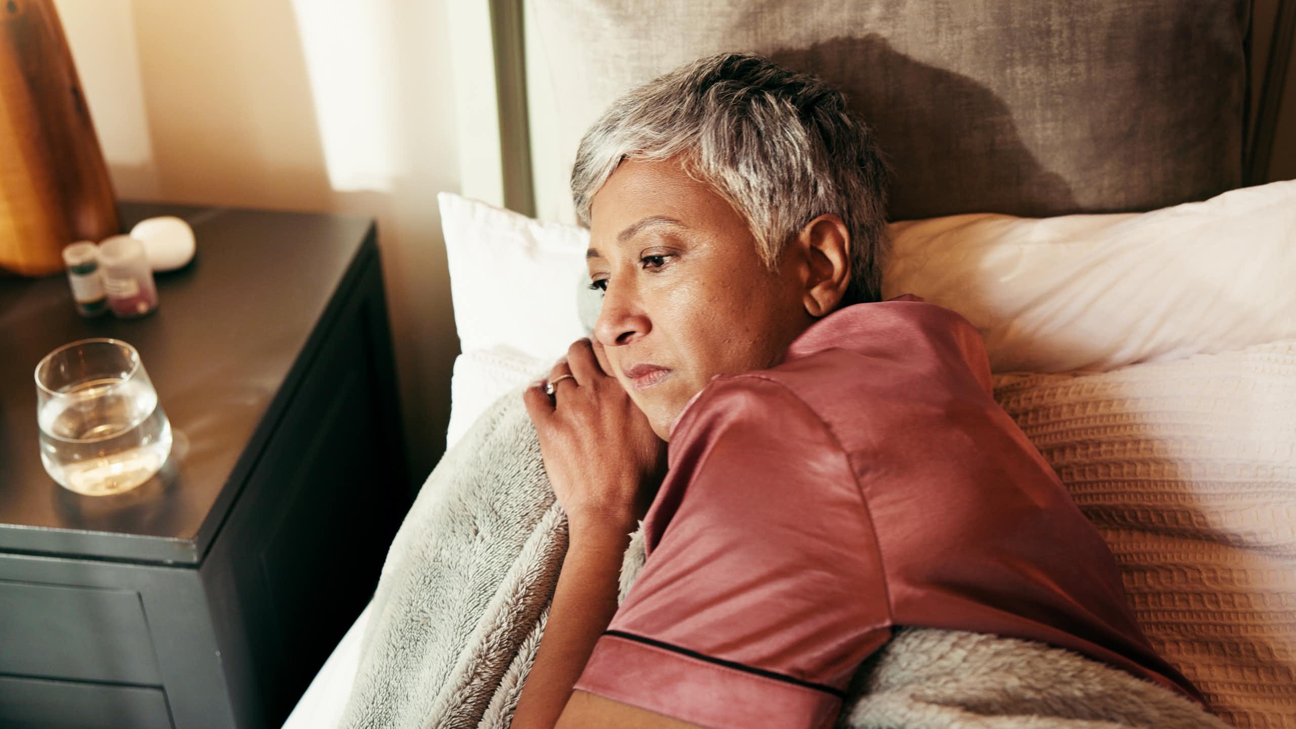 Une femme d’âge mûr est éveillée dans son lit, l’air préoccupé. Sur sa table de nuit se trouvent des boîtes de médicaments.