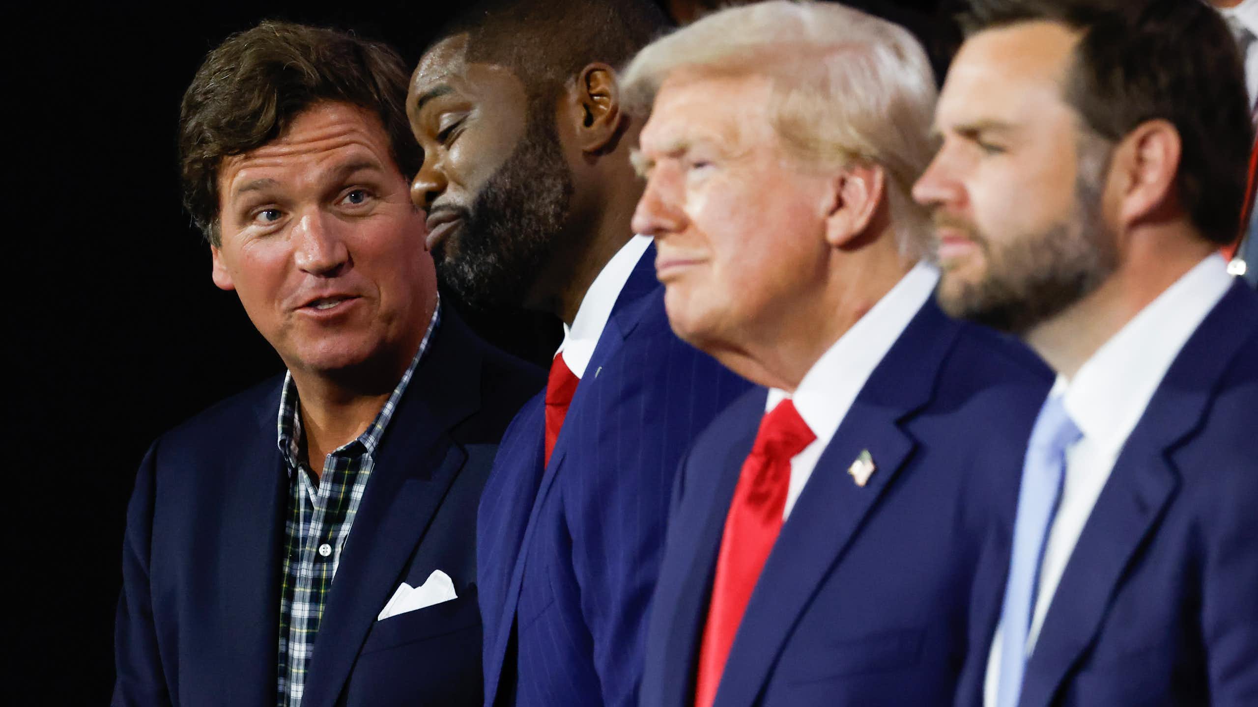 Tucker Carlson, in plaid shirt and suit jacket, with three men in suit and tie, including Donald Trump and JD Vance