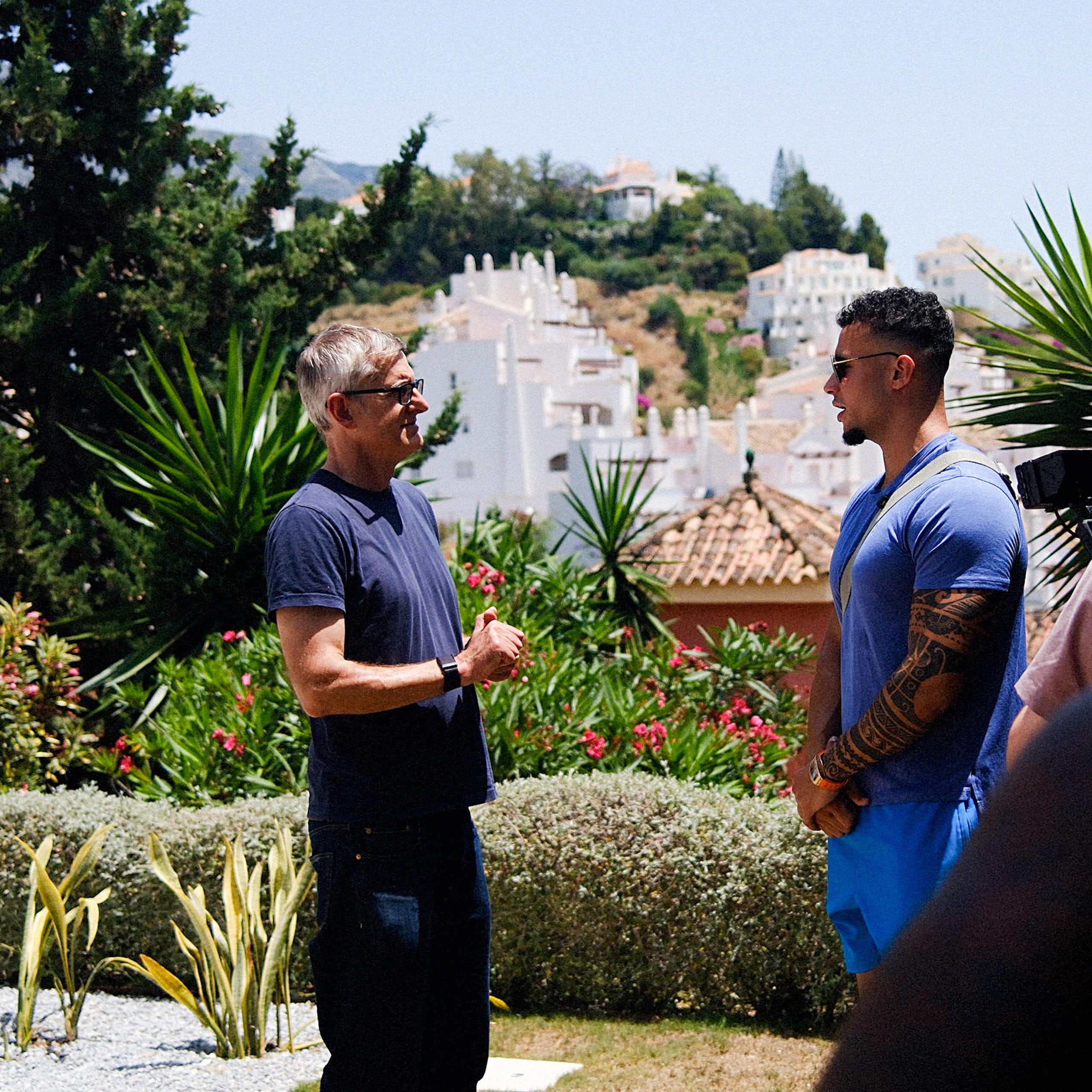A journalist interviews a muscular, tattooed man in a sunny garden. They are surrounded by a small film crew.