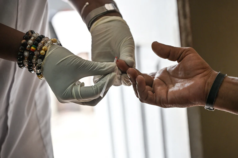 Gloved hands take a drop of blood into a glass tube from another person's fingertip