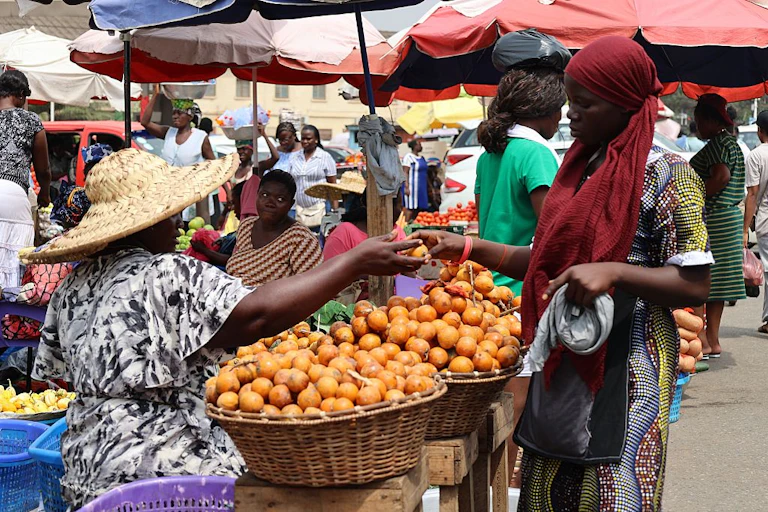 A woman at a street market under umbrellas hands fruit to another woman; baskets of the fruit are on tables