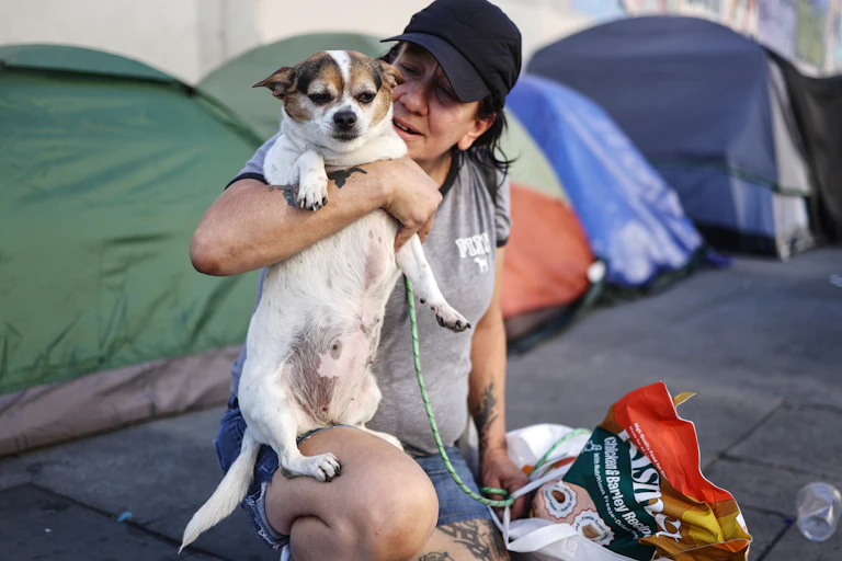 A woman holds up her dog while sitting on the sidewalk in front of a row of weathered tents.