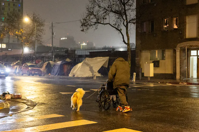 A homeless man walks a dog toward a group of tents lining a sidewalk.