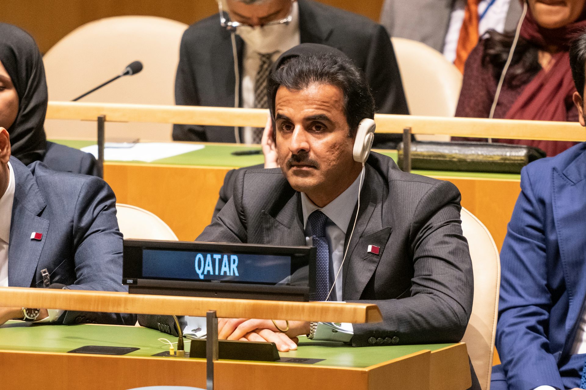 Un hombre sentado en una reunión con auriculares y el rótulo "Qatar" delante de él.