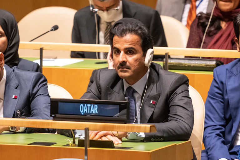 Un hombre sentado en una reunión con auriculares y el rótulo "Qatar" delante de él.